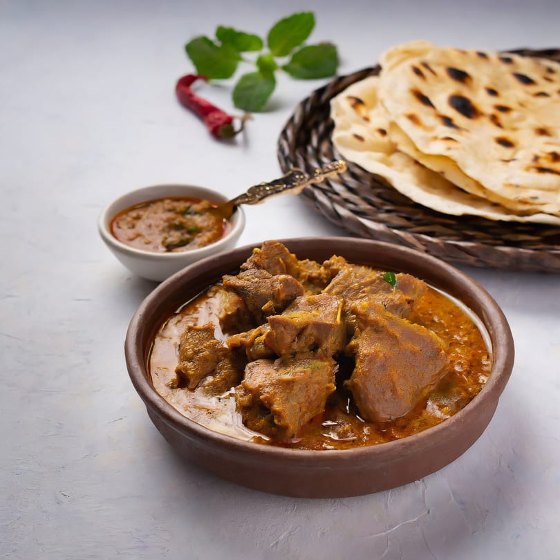 Bada Indian Tikka and Curry's lamb curry in a brown bowl, with naan bread and a side sauce.